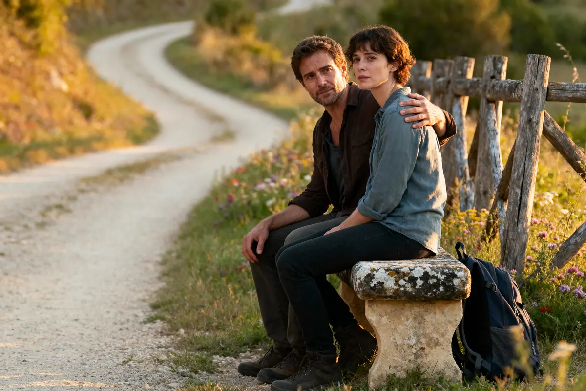 Un couple assis sur un banc en pierre, près d'un chemin rural, entouré de fleurs sauvages.