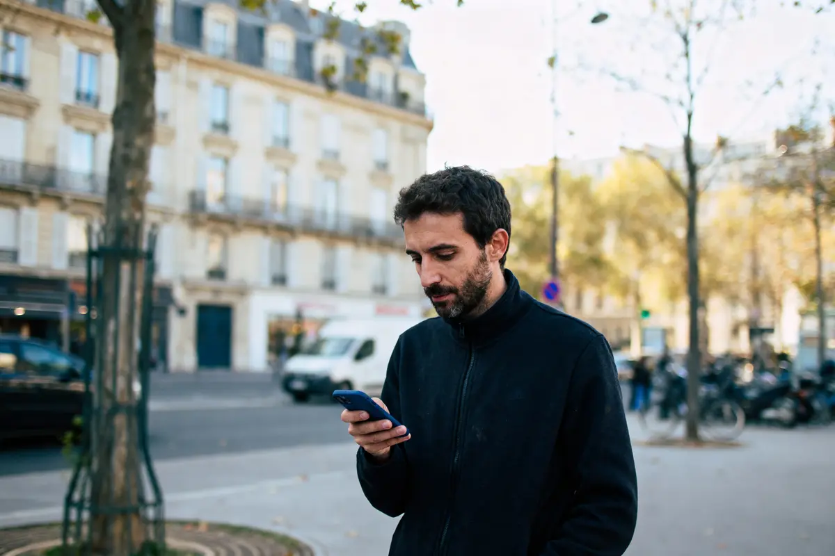 Homme lisant un smartphone dans une rue urbaine, immeubles et arbres en arrière-plan. Vêtements foncés.