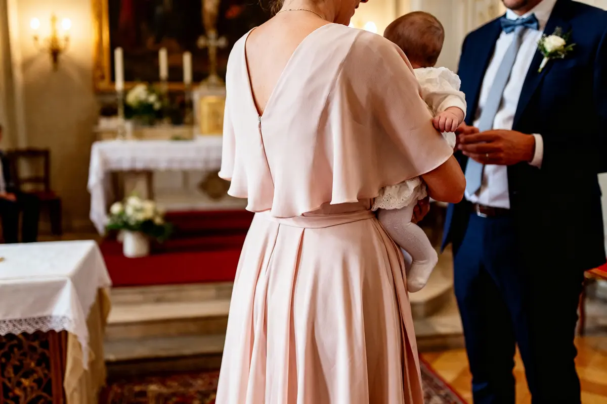 Une femme en robe rose tient un bébé, scène d'intérieur, peut-être une cérémonie religieuse.