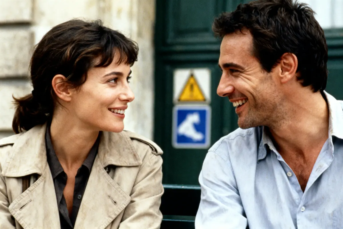 Un homme et une femme sourient, assis dehors, en train de discuter, fond de bâtiment flou.