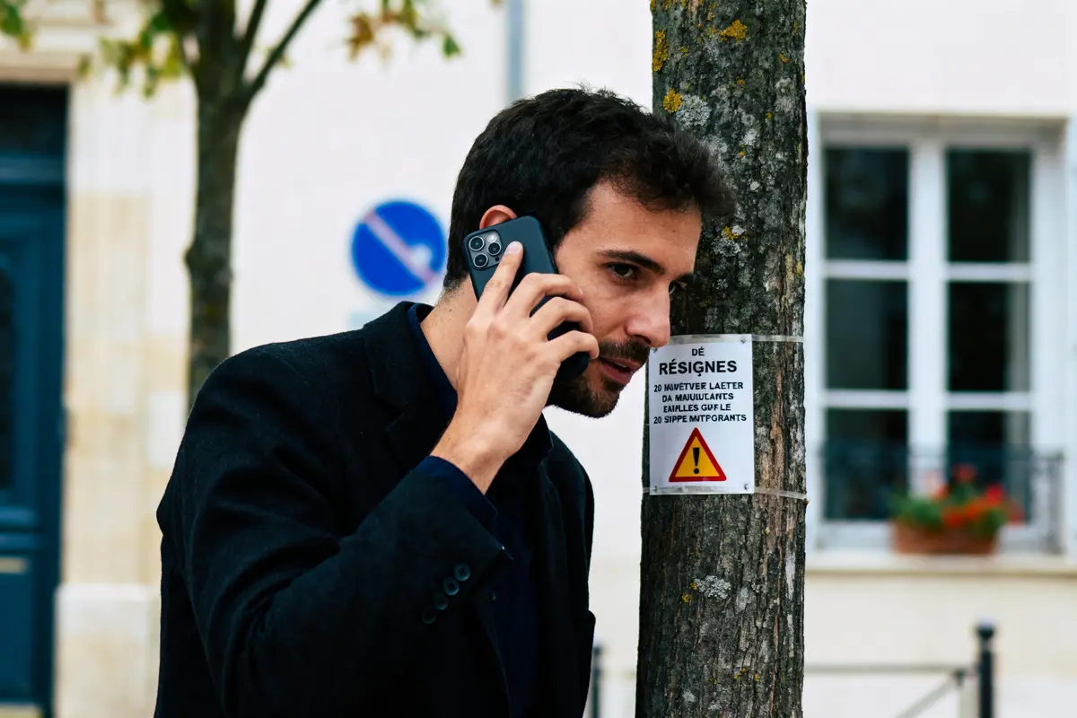 Homme parlant au téléphone, marchant près d'un arbre avec avertissement, panneau de signalisation bleu en arrière-plan.