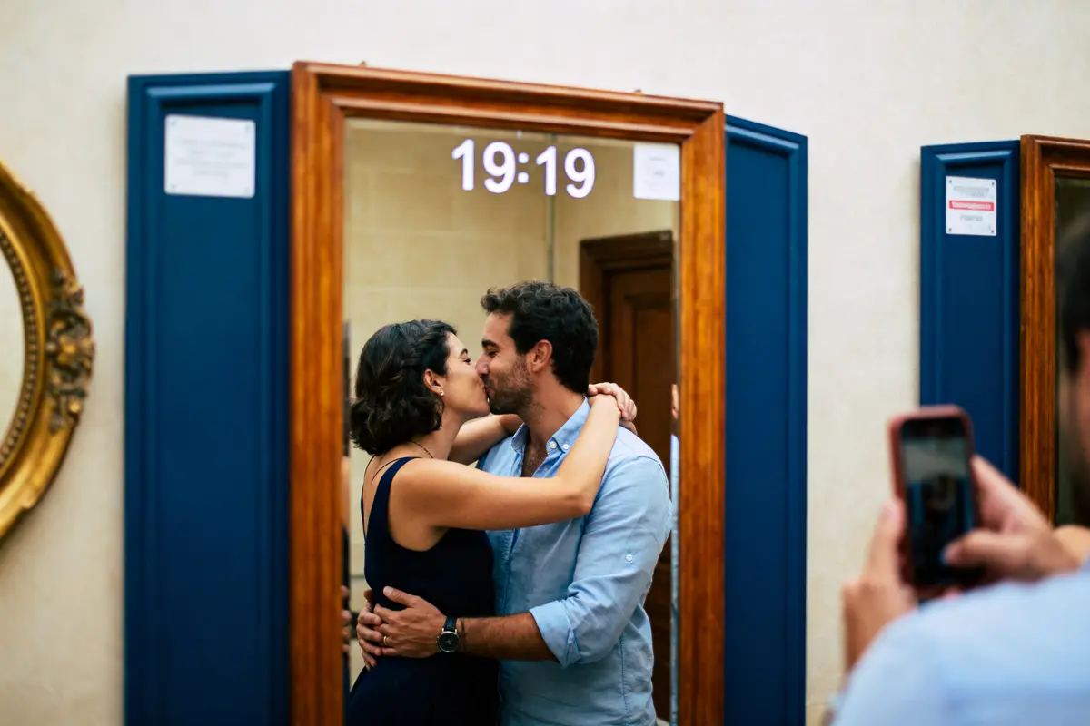 Un couple s'embrasse devant un miroir avec l'heure 19:19, un smartphone visible.