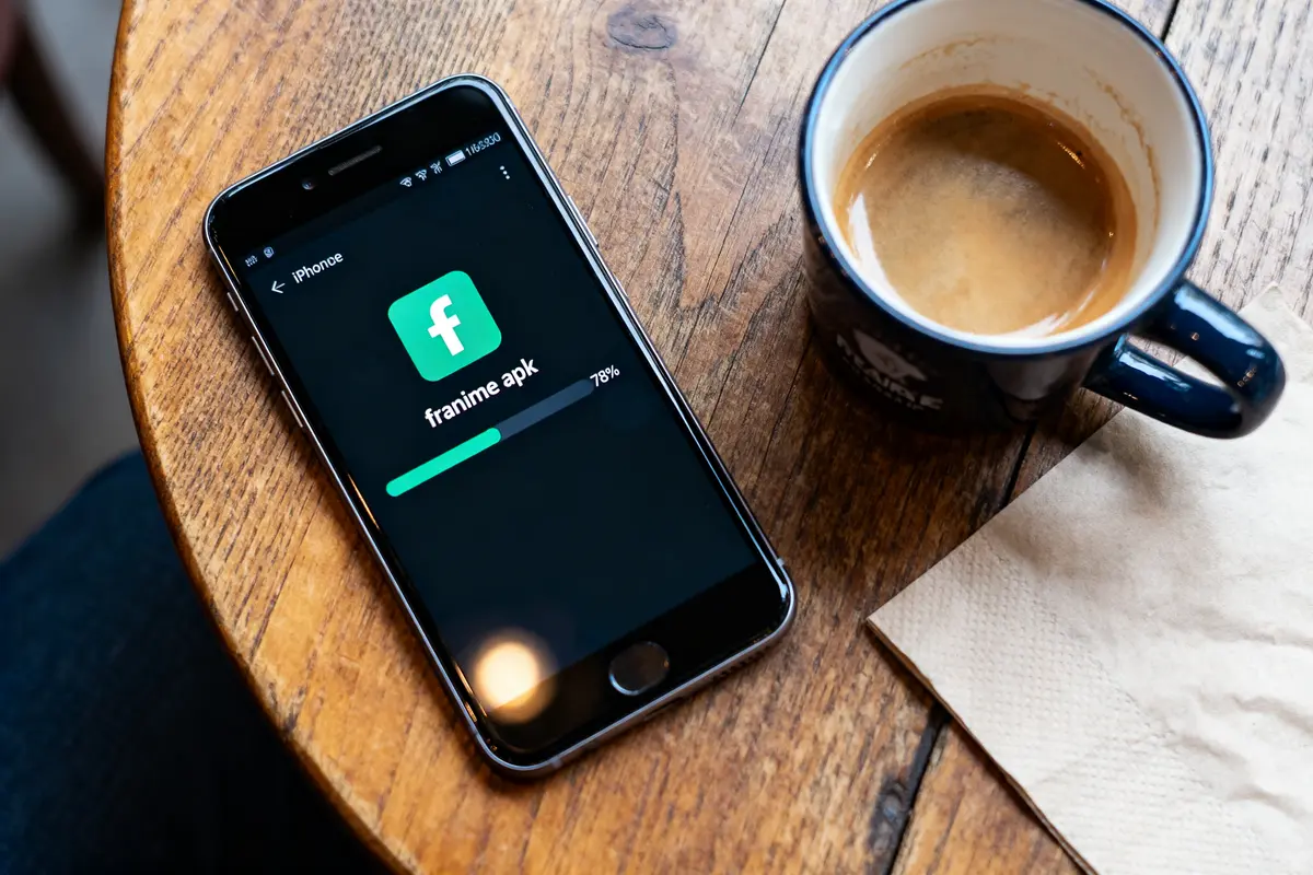 Téléphone affichant téléchargement application "franime apk", posé sur table en bois, avec tasse de café à côté.