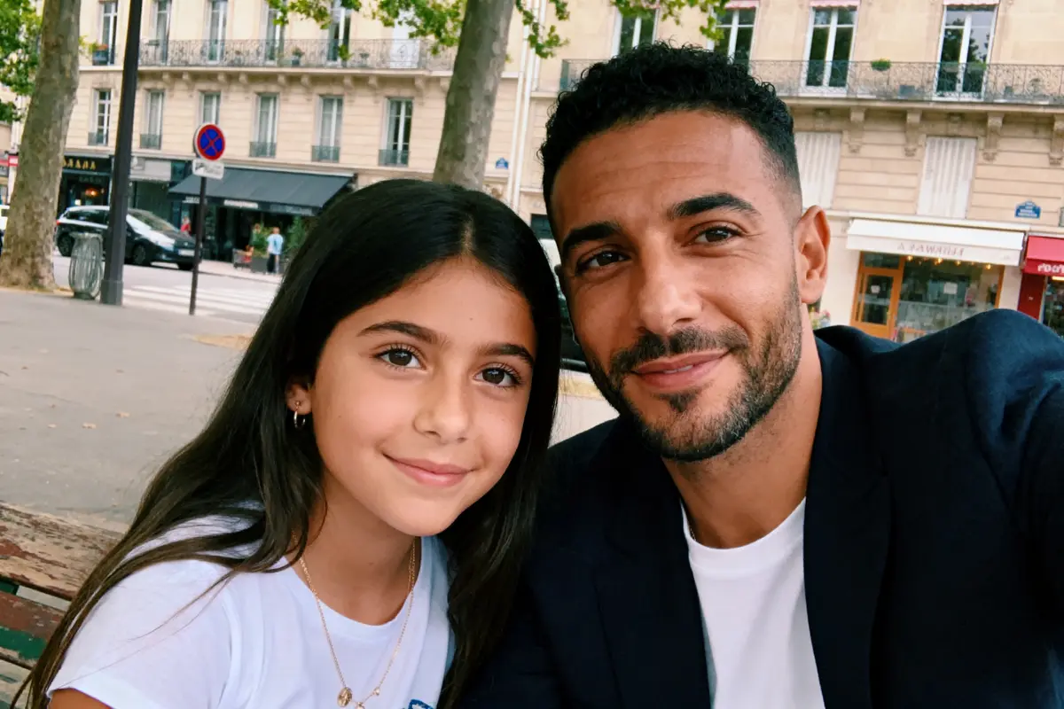 Homme et fille souriant ensemble, assis dans un parc urbain, bâtiments en arrière-plan.