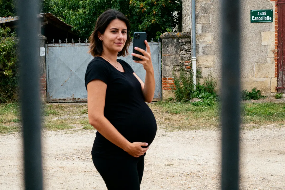 Femme enceinte en noir prenant un selfie devant portail métallique, sur chemin en terre. Pointe souriante.