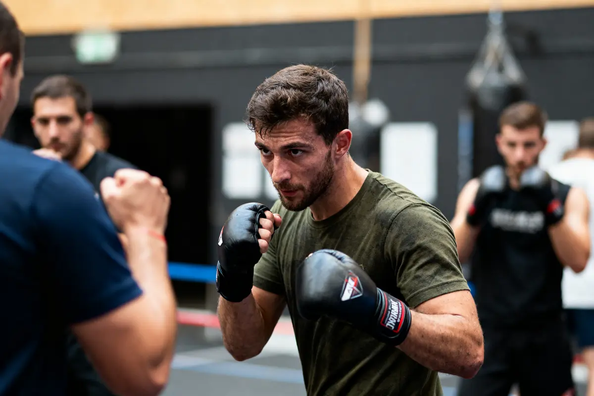 Homme en t-shirt s'entraînant à la boxe dans une salle avec d'autres participants en arrière-plan.