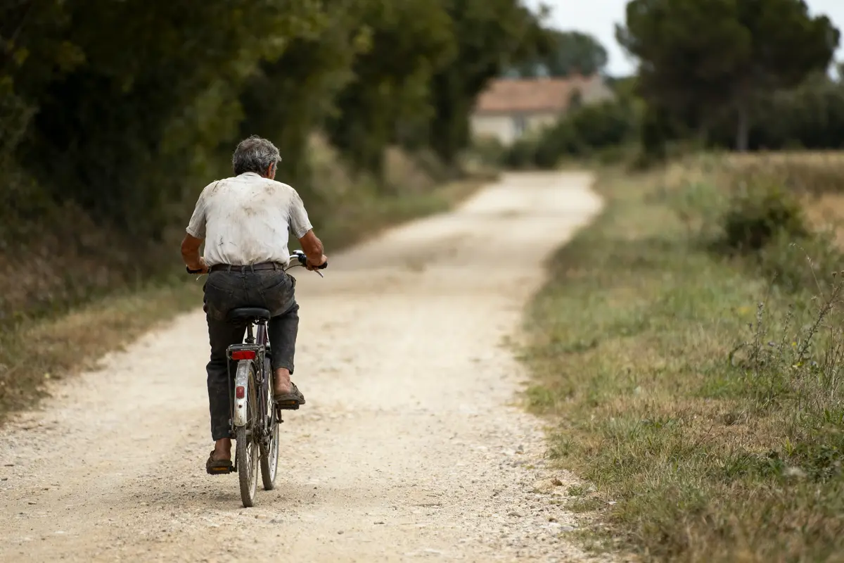 Homme âgé à vélo sur un chemin rural entouré d'arbres et de verdure.