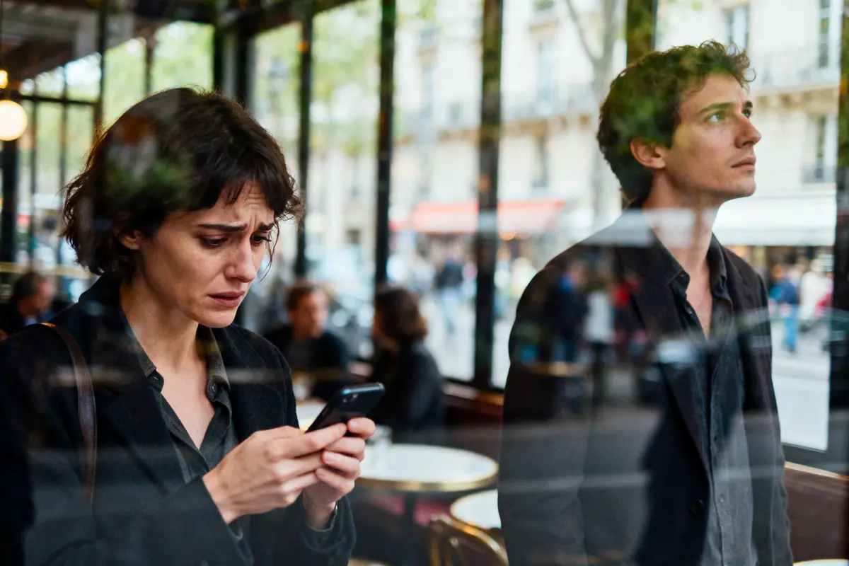 Personnes réfléchissant dans un café, femme tenant un téléphone, environnement urbain visible à travers la vitre.