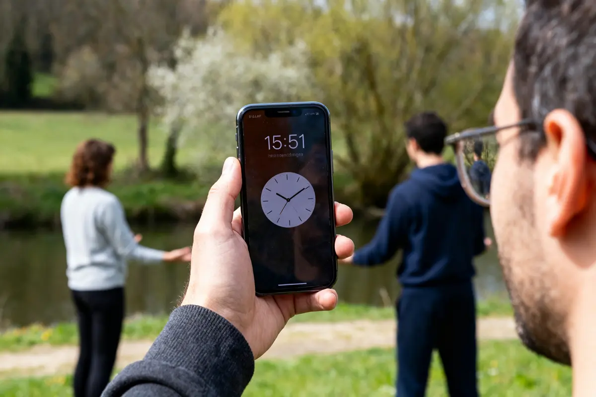 Une main tient un smartphone montrant l'heure, deux personnes méditant près d'une rivière.