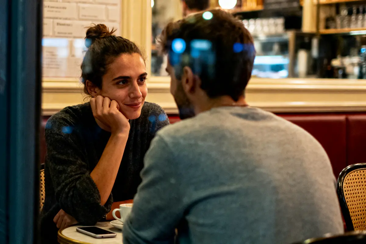 Deux personnes discutant dans un café, une femme souriante regardant un homme assis en face d'elle.