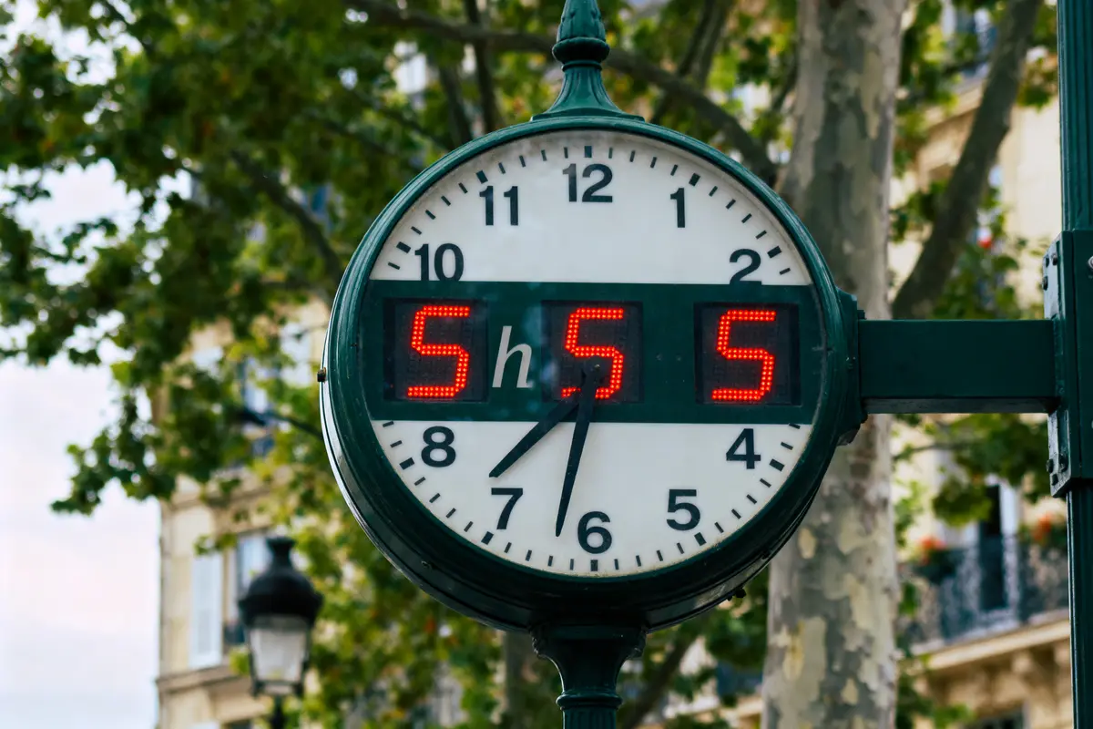 Horloge analogique à l'extérieur affichant 5h55 avec chiffres numériques rouges. Arrière-plan arboré.