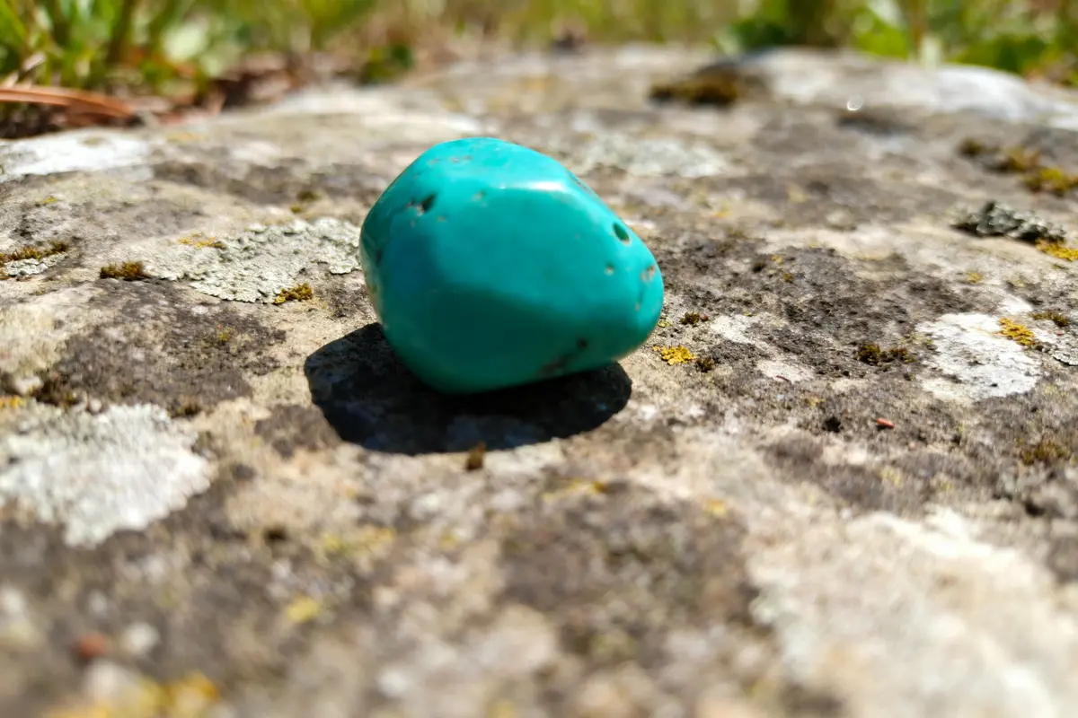 Pierre turquoise posée sur une surface rocheuse en extérieur, avec herbe floue en arrière-plan.